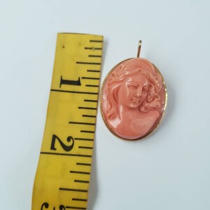 May include: A coral-colored cameo pendant with a woman's profile carved in relief. The oval pendant is set in a gold-colored frame. The pendant is approximately 1.75 inches in length, as measured by a yellow ruler.