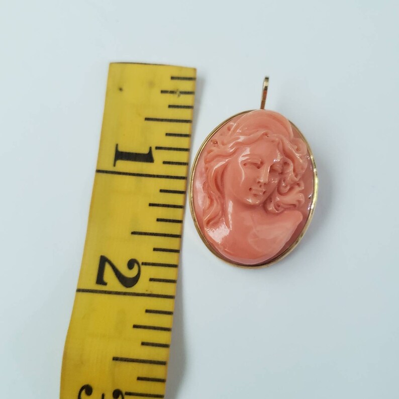 May include: A coral-colored cameo pendant with a woman's profile carved in relief. The oval pendant is set in a gold-colored frame. The pendant is approximately 1.75 inches in length, as measured by a yellow ruler.