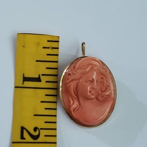 May include: Oval pendant with a carved coral cameo of a woman's profile, set in a gold frame. The pendant is approximately 1.5 inches in length. The cameo features detailed carving and a warm coral hue, with a gold bail for hanging.