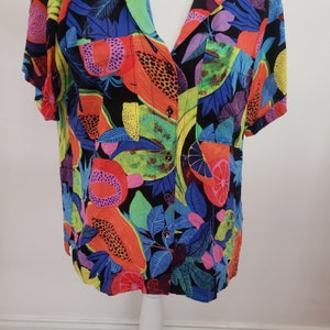 May include: A colorful short-sleeved shirt with a tropical fruit print. The shirt features a variety of fruits, including pineapples, oranges, and mangoes, in shades of yellow, orange, red, and green. The background is a dark blue with a black pattern.