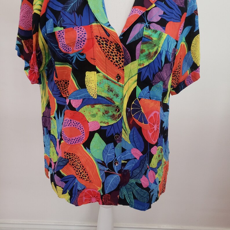 May include: A colorful short-sleeved shirt with a tropical fruit print. The shirt features a variety of fruits, including pineapples, oranges, and mangoes, in shades of yellow, orange, red, and green. The background is a dark blue with a black pattern.