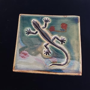 May include: A ceramic tile with a green and brown glaze features a black lizard in relief. The lizard is facing right and has a long tail.