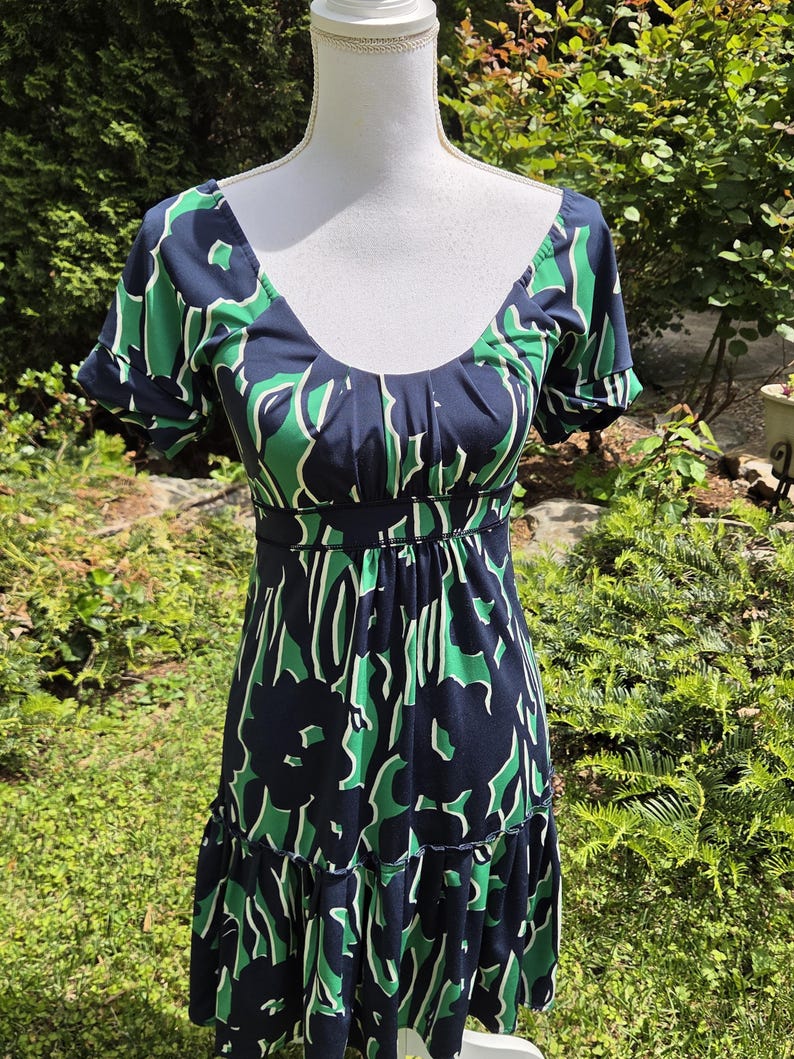Puede incluir: Vestido con estampado floral azul marino y verde, con escote en forma de U y mangas cortas. El vestido tiene un corpi&ntilde;o ajustado y una falda escalonada. El vestido se exhibe en un maniqu&iacute; blanco en un entorno al aire libre.