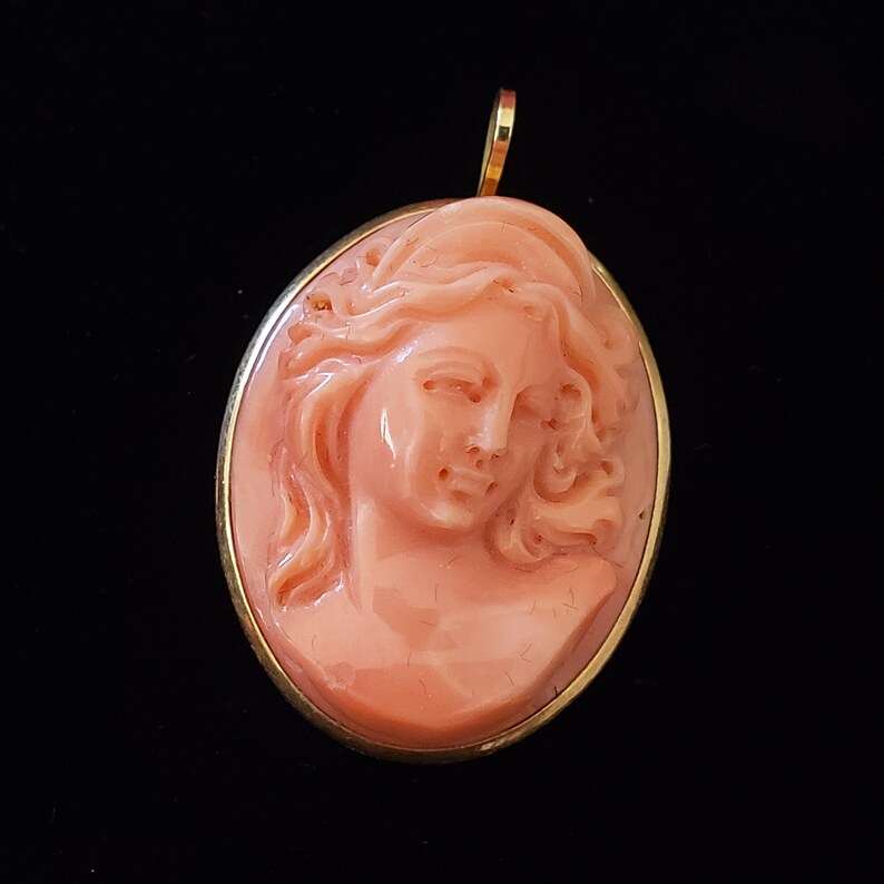 May include: Oval pendant featuring a carved coral cameo of a woman's profile. The cameo is a light peach color, set in a gold-toned frame with a small loop at the top for a chain. The background is black.