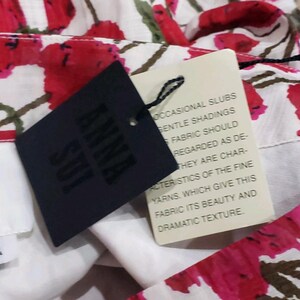 May include: Close-up of a floral fabric with red and white flowers and green leaves. A black tag with the brand name "ANNA SUI" is attached. A cream-colored tag with text about the fabric's characteristics is also visible.