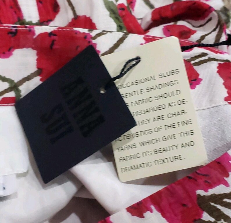 May include: Close-up of a floral fabric with red and white flowers and green leaves. A black tag with the brand name "ANNA SUI" is attached. A cream-colored tag with text about the fabric's characteristics is also visible.