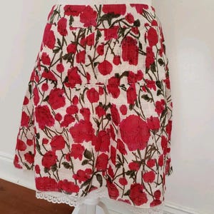 May include: A-line skirt with a floral pattern. The skirt features a white background with large red flowers and green stems. The hemline is trimmed with white lace.