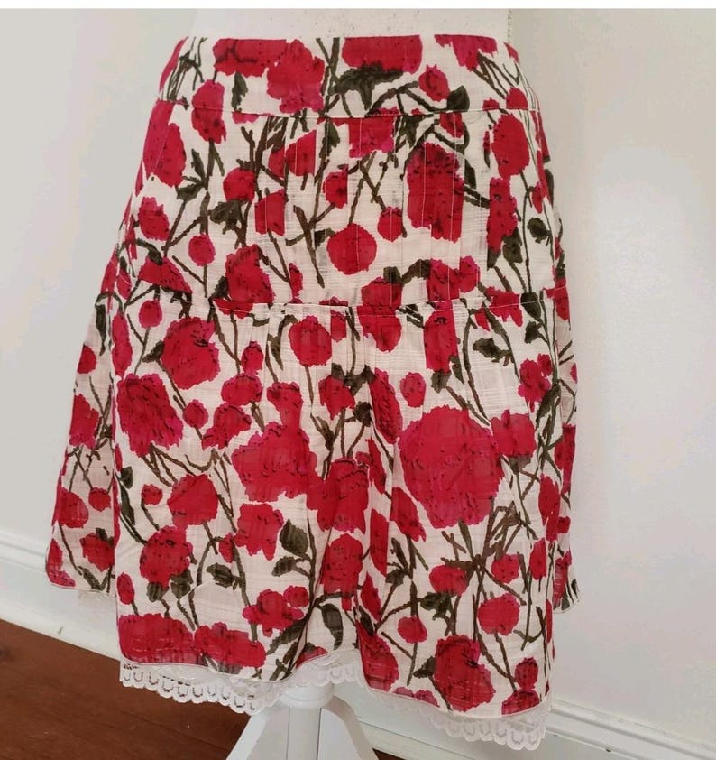May include: A-line skirt with a floral pattern. The skirt features a white background with large red flowers and green stems. The hemline is trimmed with white lace.