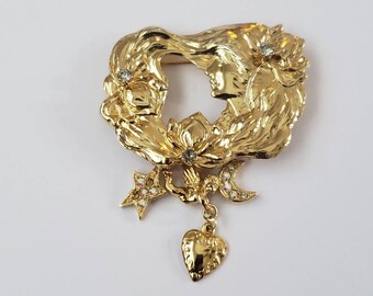 May include: A gold-tone brooch featuring a stylized image of two women's faces with hair flowing around them. The brooch has a heart-shaped pendant with a star and crescent moon charm hanging from it.