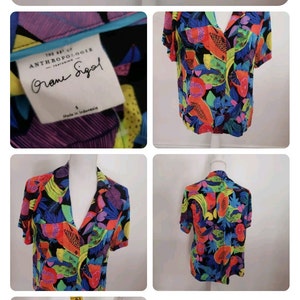 May include: A black short-sleeved button-up shirt with a colorful tropical fruit print. The shirt has a collar and a button-up front. The print features papaya, bananas, and other tropical fruits in shades of red, orange, yellow, green, blue, and purple. The shirt is made by Anthropologie and features the text "The Art of Anthropologie featuring  " in a white font. The shirt is size small and made in Indonesia.