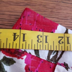 May include: Close-up of a floral fabric with a yellow measuring tape. The fabric features a pattern of red and pink flowers with green leaves on a white background. The measuring tape shows measurements from 12 to 15 inches.