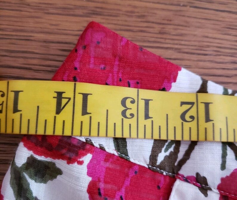 May include: Close-up of a floral fabric with a yellow measuring tape. The fabric features a pattern of red and pink flowers with green leaves on a white background. The measuring tape shows measurements from 12 to 15 inches.
