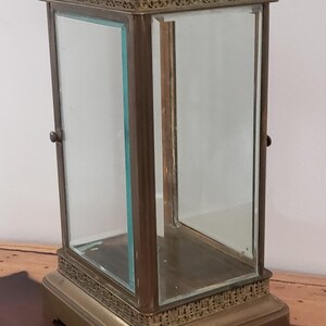 K&ouml;nnte beinhalten: Eine Vitrine aus Messing mit Glasseiten und einer Glasplatte. Die Vitrine hat einen dekorativen Rand um die Ober- und Unterseite. Die Vitrine steht auf einer Holzoberfl&auml;che.
