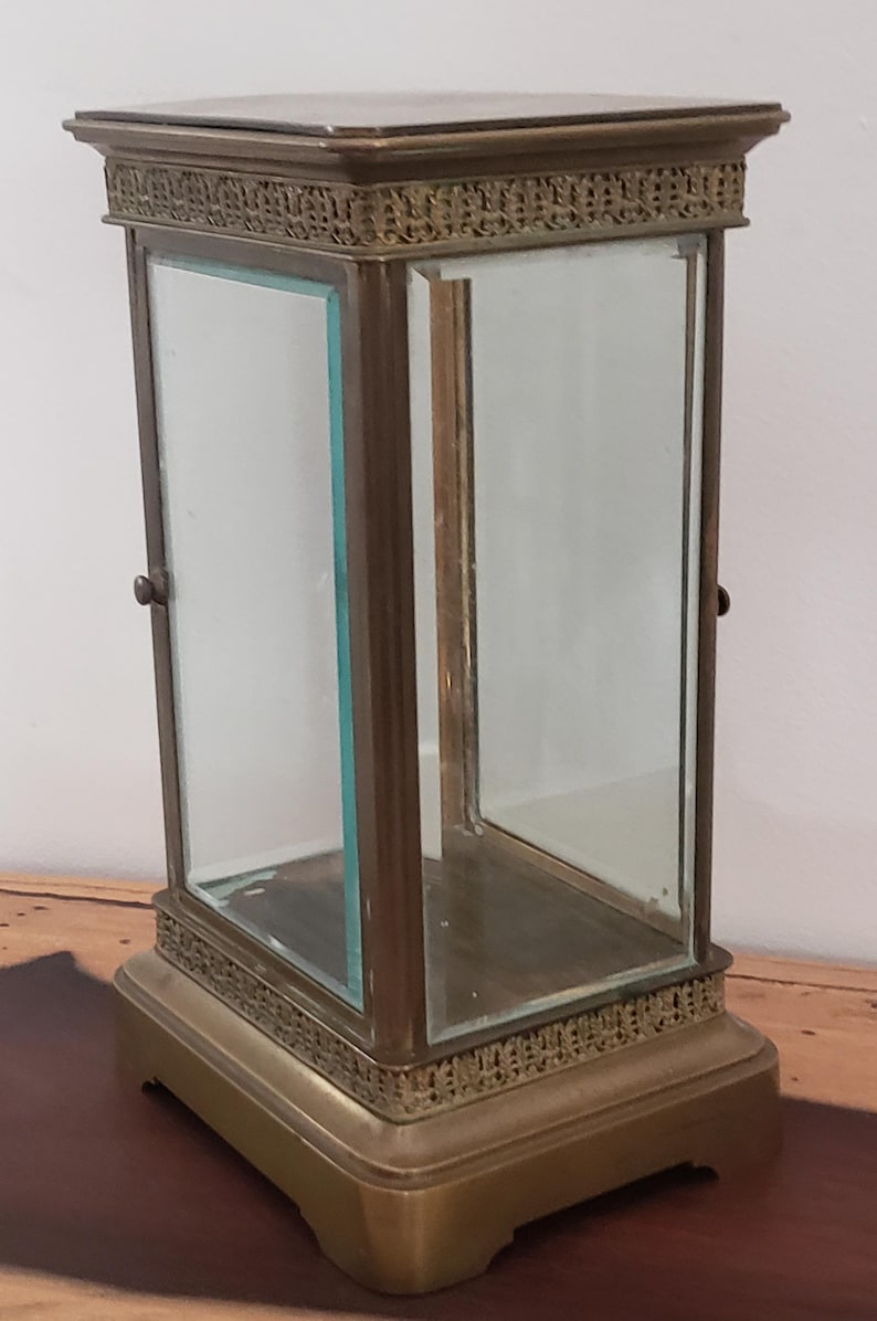 K&ouml;nnte beinhalten: Eine Vitrine aus Messing mit Glasseiten und einer Glasplatte. Die Vitrine hat einen dekorativen Rand um die Ober- und Unterseite. Die Vitrine steht auf einer Holzoberfl&auml;che.