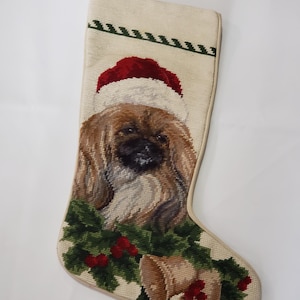 May include: Cream-colored Christmas stocking with a detailed needlepoint design. Features a dog wearing a Santa hat, surrounded by holly, berries, and bells. The top edge has a green zig-zag pattern. A festive holiday decoration.