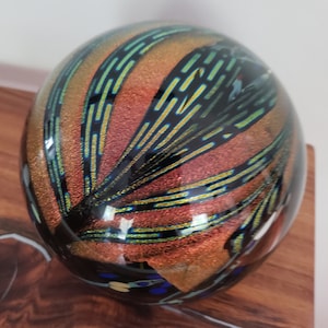 Könnte beinhalten: Eine runde, irisierende Glaskugel mit einem abstrakten Design in Schwarz, Grün und Gold. Die Kugel liegt auf einer Holzoberfläche.
