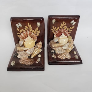 May include: A pair of wooden bookends with a seashell design. The bookends are brown and feature a variety of seashells in shades of white, brown, and pink.