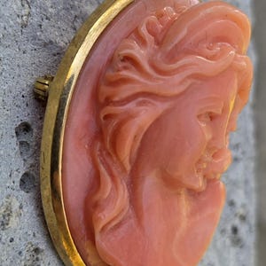 May include: Oval pendant with a carved cameo of a woman's profile in a light coral color. The cameo is set in a gold-colored frame with a loop at the top for a chain. The pendant is likely vintage or antique.