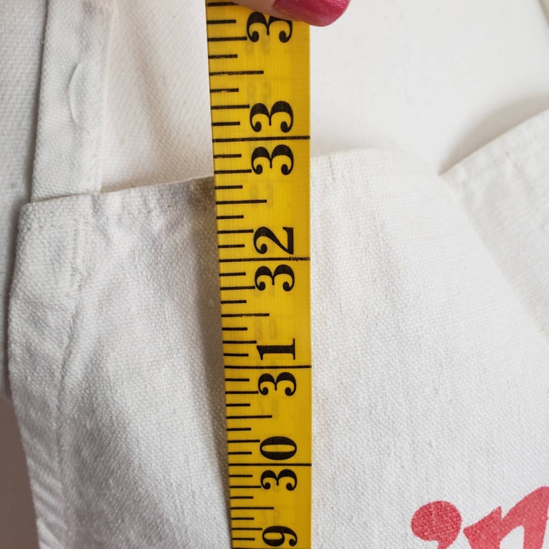 May include: A white canvas tote bag with a yellow measuring tape showing a measurement of 33 inches. The tote bag has a red graphic on the front.