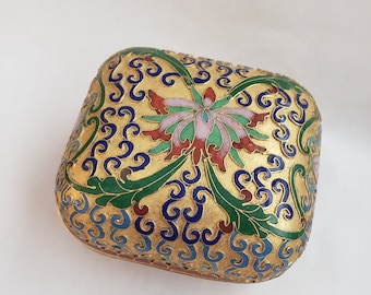 May include: A small, square, gold-colored box with a floral design. The box is decorated with blue, green, pink, and red enamel. The design features a large flower in the center, surrounded by smaller flowers and leaves.