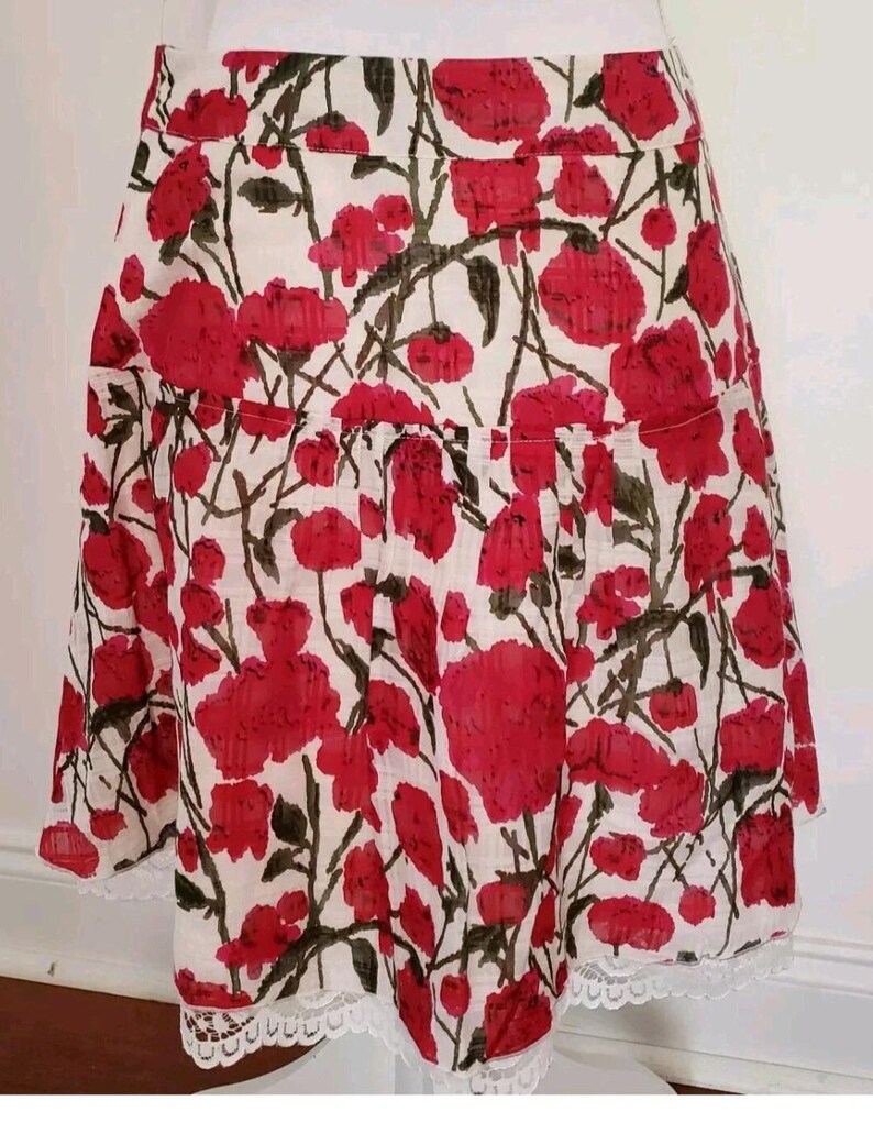 May include: A white skirt with a red floral pattern. The skirt features a ruffled hem and a white lace trim along the bottom edge. The floral design includes red flowers and green leaves.