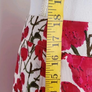 May include: A floral skirt with a white background and red flower pattern. The skirt is being measured with a yellow measuring tape. The measurement is between 15 and 19 inches.