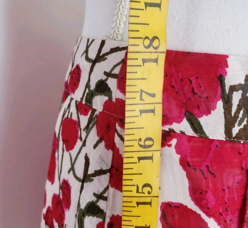 May include: A floral skirt with a white background and red flower pattern. The skirt is being measured with a yellow measuring tape. The measurement is between 15 and 19 inches.