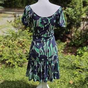 Puede incluir: Vestido azul marino con estampado floral verde y blanco. El vestido tiene mangas cortas, cintura entallada y bajo con volantes. Se exhibe en un maniqu&iacute; blanco en un jard&iacute;n.