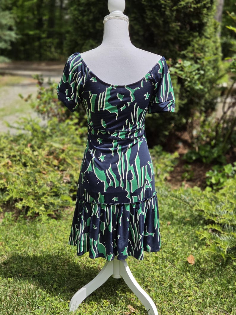 Puede incluir: Vestido azul marino con estampado floral verde y blanco. El vestido tiene mangas cortas, cintura entallada y bajo con volantes. Se exhibe en un maniqu&iacute; blanco en un jard&iacute;n.