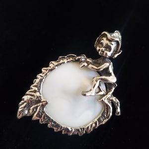 May include: A silver charm featuring a small fairy sitting on a white pearl-like disc surrounded by a leafy design.