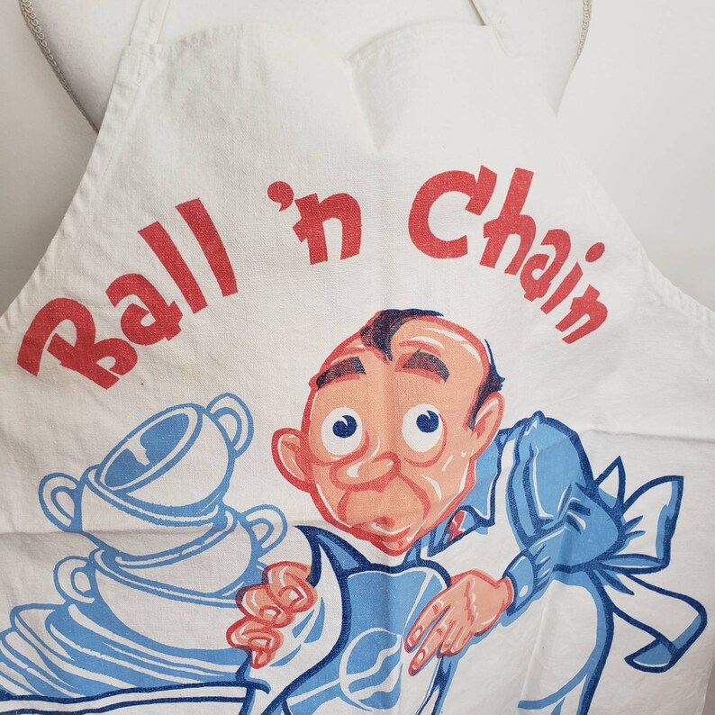 May include: White kitchen apron with blue and white dishes and a cartoon man with red hair and blue shirt. The text "Ball 'n Chain" is printed in red on the apron.