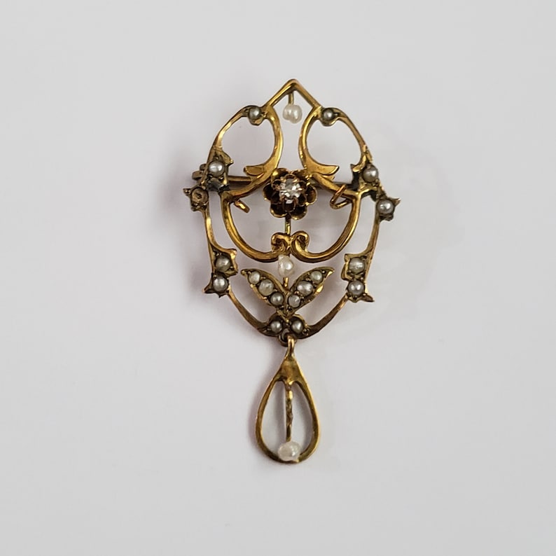 May include: A gold pendant brooch with a teardrop design. The pendant is adorned with small pearls and a single diamond in the center.