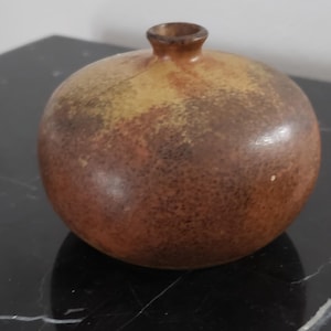May include: A small, round ceramic vase with a narrow neck. The vase has a speckled, earthy color palette of brown, tan, and rust. The spherical shape and muted tones suggest a rustic or minimalist aesthetic.