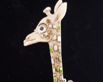 May include: A silver giraffe brooch with green and white enamel spots. The giraffe's head is turned to the side and has a large, white eye.