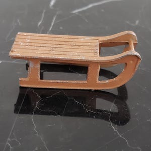 May include: A small, vintage toy sled in a warm, muted orange hue. The sled features a slatted seat and curved runners, showing signs of wear. The design is reminiscent of classic winter fun.