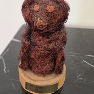 May include: A handcrafted wire sculpture of a teddy bear. The bear is made of reddish-brown wire and sits on a wooden base with a metal plaque that reads "WIRE ORIGINALS by RAYMOND". The bear's eyes and nose are also made of wire.