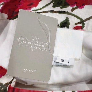 May include: Close-up of a floral print garment with a tag from Anthropologie. The tag is gray with the brand name and the word "apparel" printed on it. The garment has a white background with red and green floral patterns.