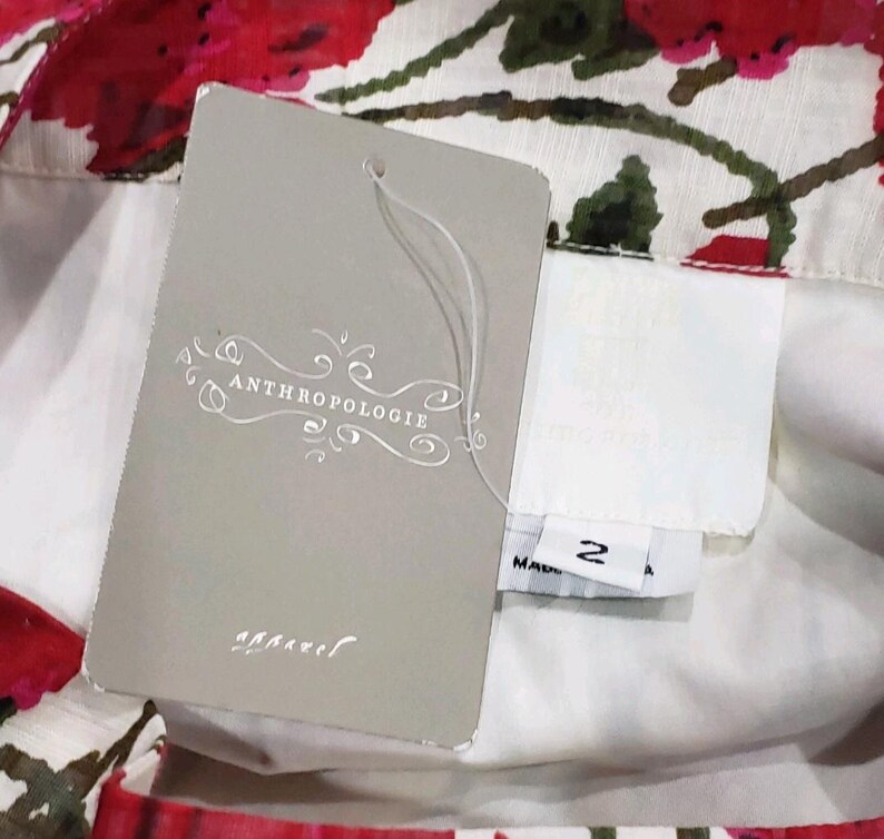 May include: Close-up of a floral print garment with a tag from Anthropologie. The tag is gray with the brand name and the word "apparel" printed on it. The garment has a white background with red and green floral patterns.