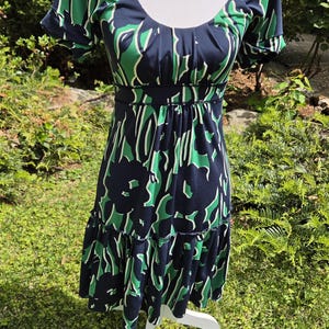 Puede incluir: Vestido azul marino con estampado floral verde y blanco. El vestido tiene mangas cortas, cuello redondo y cintura entallada. La falda tiene un dobladillo con volantes escalonados. El vestido se exhibe en un maniqu&iacute; blanco.
