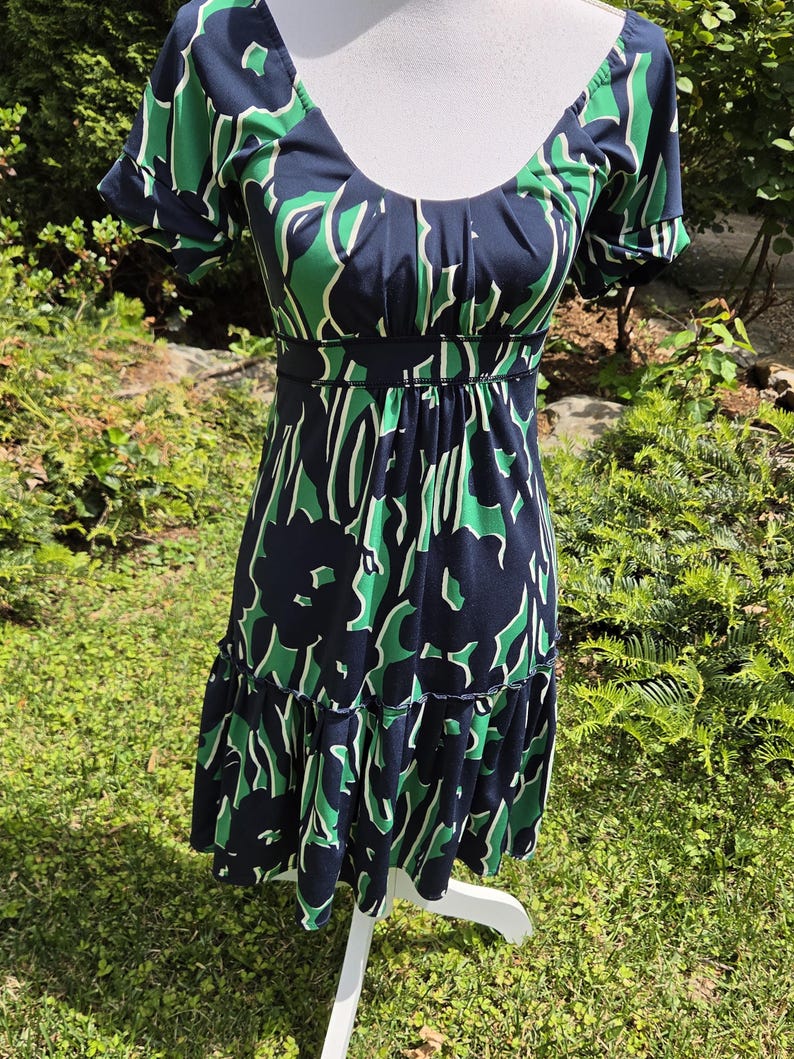 Puede incluir: Vestido azul marino con estampado floral verde y blanco. El vestido tiene mangas cortas, cuello redondo y cintura entallada. La falda tiene un dobladillo con volantes escalonados. El vestido se exhibe en un maniqu&iacute; blanco.