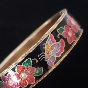 May include: A black enamel bangle bracelet with a gold trim and colorful floral and butterfly design. The flowers are red, pink, and white with green leaves. The butterfly is purple, orange, and pink.