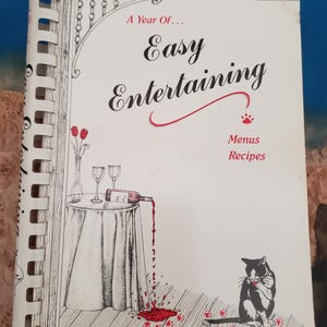May include: A spiral-bound cookbook titled "A Year Of... Easy Entertaining." The cover features a black and white illustration of a table with wine glasses and a bottle pouring wine, a cat, and the words "Menus Recipes."