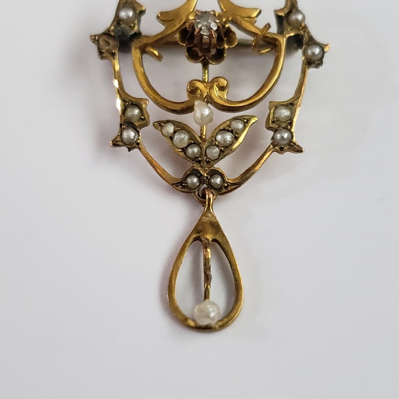 May include: A gold pendant with a teardrop-shaped design, adorned with small white pearls and a single larger pearl at the bottom. The pendant features intricate floral motifs and a small diamond in the center.