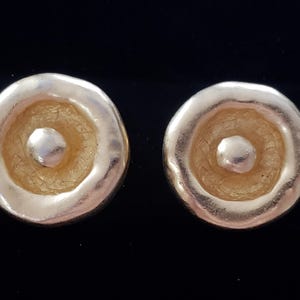 May include: A pair of gold-toned earrings with a circular design. Each earring features a raised, textured center circle with a smaller, smooth circle in the center.