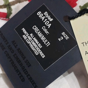 May include: A close-up of a black clothing tag with the text "Style# 69A10A, Color: CREAM/MULTI, Size 2". The tag also includes the text "ALLANNA SUI PRINTS AND EMBROIDERIES ARE COPYRIGHTED".