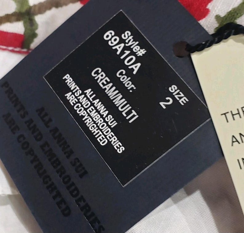 May include: A close-up of a black clothing tag with the text "Style# 69A10A, Color: CREAM/MULTI, Size 2". The tag also includes the text "ALLANNA SUI PRINTS AND EMBROIDERIES ARE COPYRIGHTED".