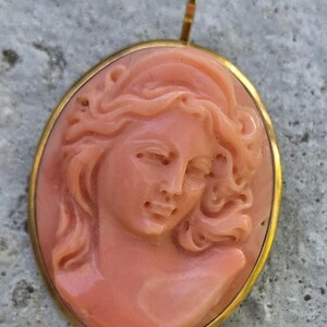 May include: A coral-colored cameo pendant featuring a carved profile of a woman with flowing hair. The oval pendant is set in a gold-colored frame with a small loop at the top for a chain.