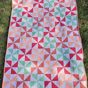 May include: A rectangular quilt featuring a repeating pinwheel pattern composed of triangular fabric segments. The design incorporates solid red, light green, and peach-colored triangles interspersed with white fabric printed with small floral patterns.
