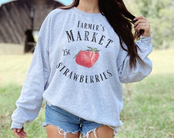 May include: A gray sweatshirt with the text "Farmer's Market 25c Strawberries" and a red strawberry graphic. The model is wearing the sweatshirt with a pair of denim shorts and white cowboy boots.