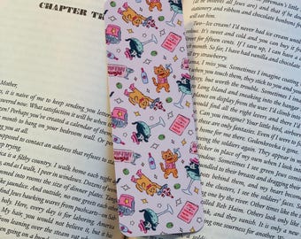 May include: Pink bookmark with a repeating pattern of cartoon cats, cocktails, and other whimsical elements. The bookmark is placed in an open book, with text visible on the pages. The bookmark is rectangular and has rounded corners.